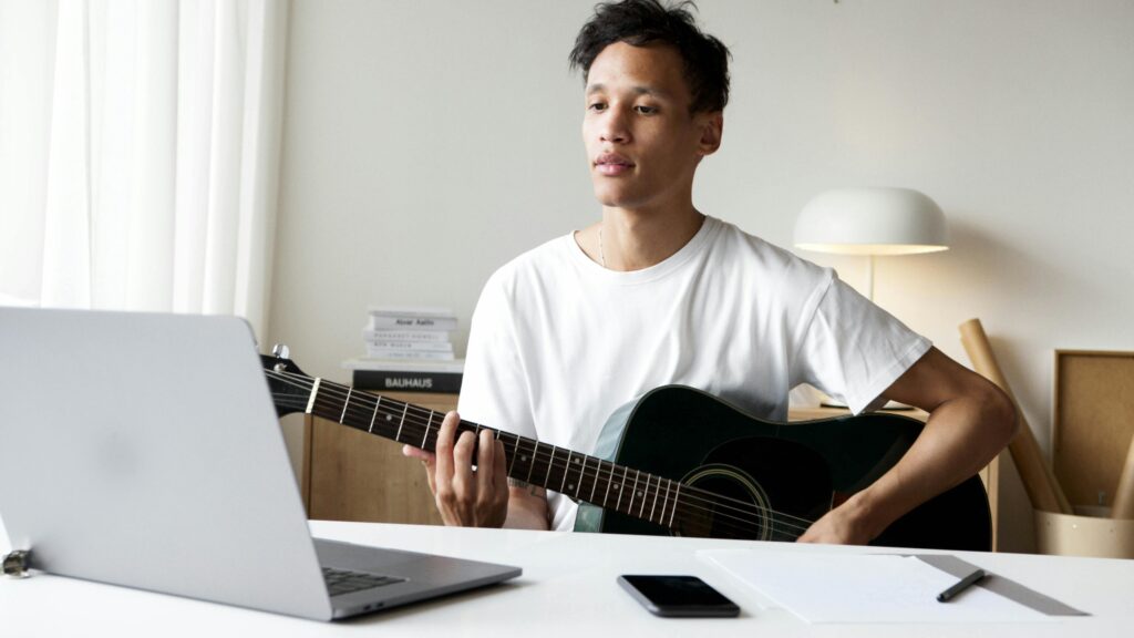 Man playing an acoustic guitar while using a laptop in a well-lit room, ideal for music creativity concepts.