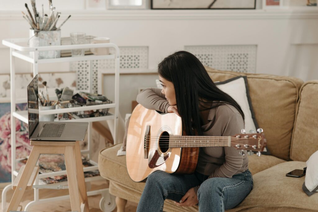 A woman sitting on a sofa with a guitar, learning music online through a laptop.