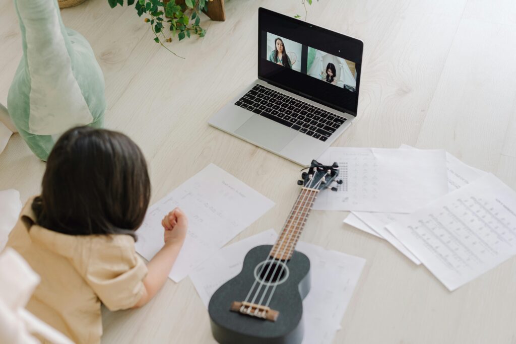 A child participating in an online music lesson with a ukulele, music sheets, and a laptop showing a video call.